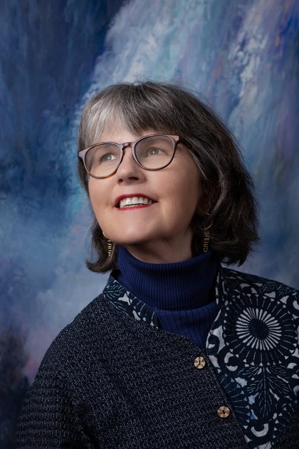 Pamela Belyea, in glasses and a dark patterned jacket, looking up and smiling, against one of Gary Faigin’s blue paintings.